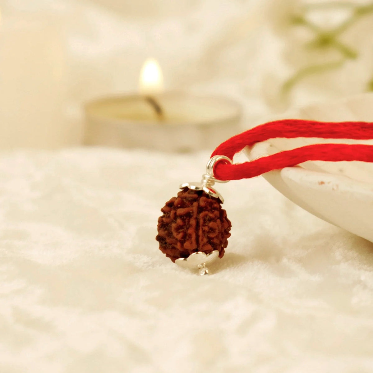 VASTUKOSHA 3 Mukhi Nepal Rudraksha bead with silver cap on a red thread, set against a soft, out-of-focus candle background.