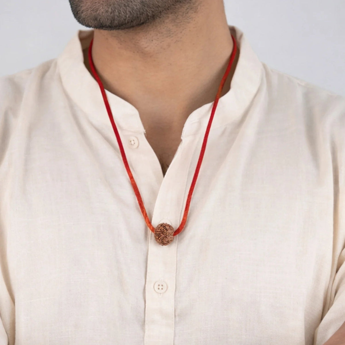 Man wearing a VASTUKOSHA 7 Mukhi Nepal origin Rudraksha bead on a simple red thread necklace against a white background.
