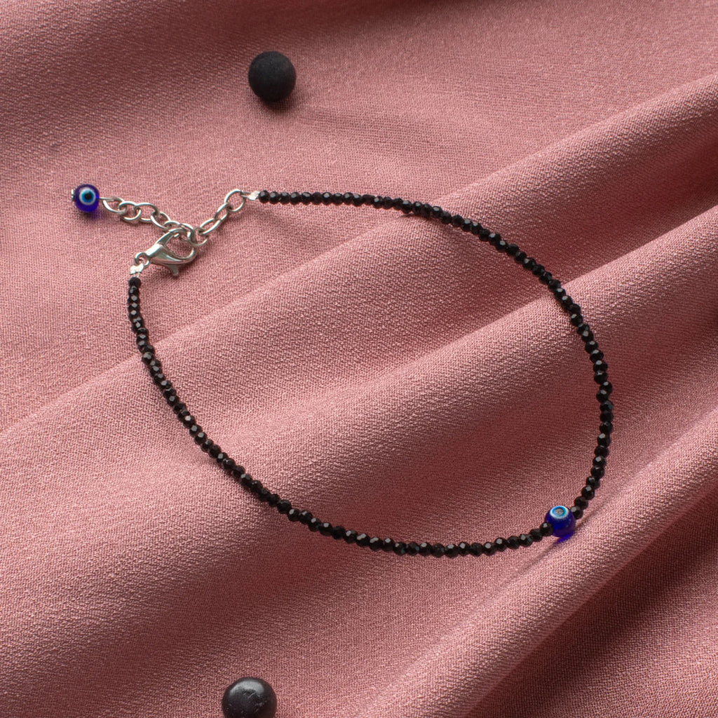 VASTUKOSHA black tourmaline beaded anklet with a blue evil eye charm on a soft pink fabric background.