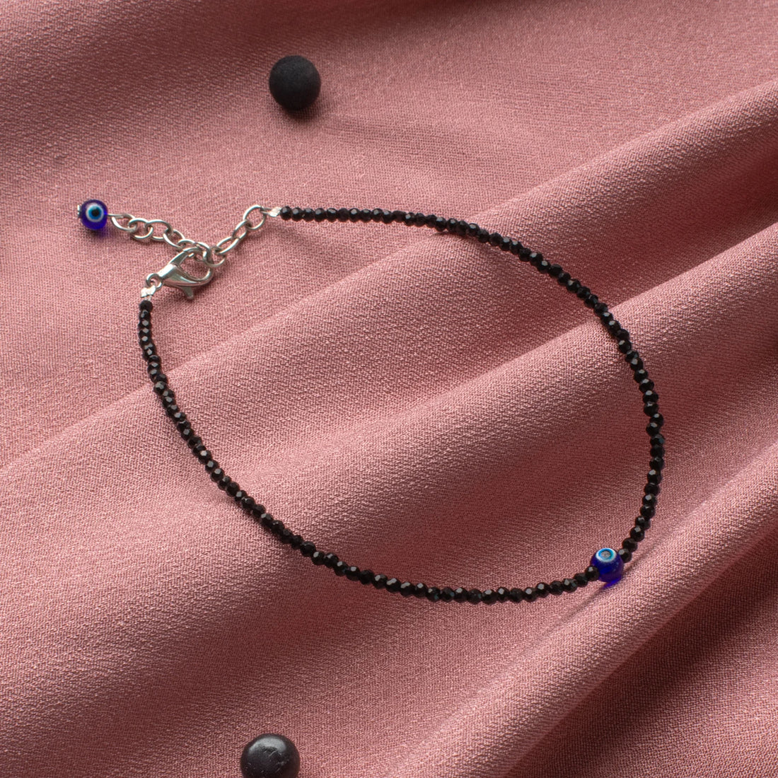 VASTUKOSHA black tourmaline beaded anklet with a blue evil eye charm on a soft pink fabric background.