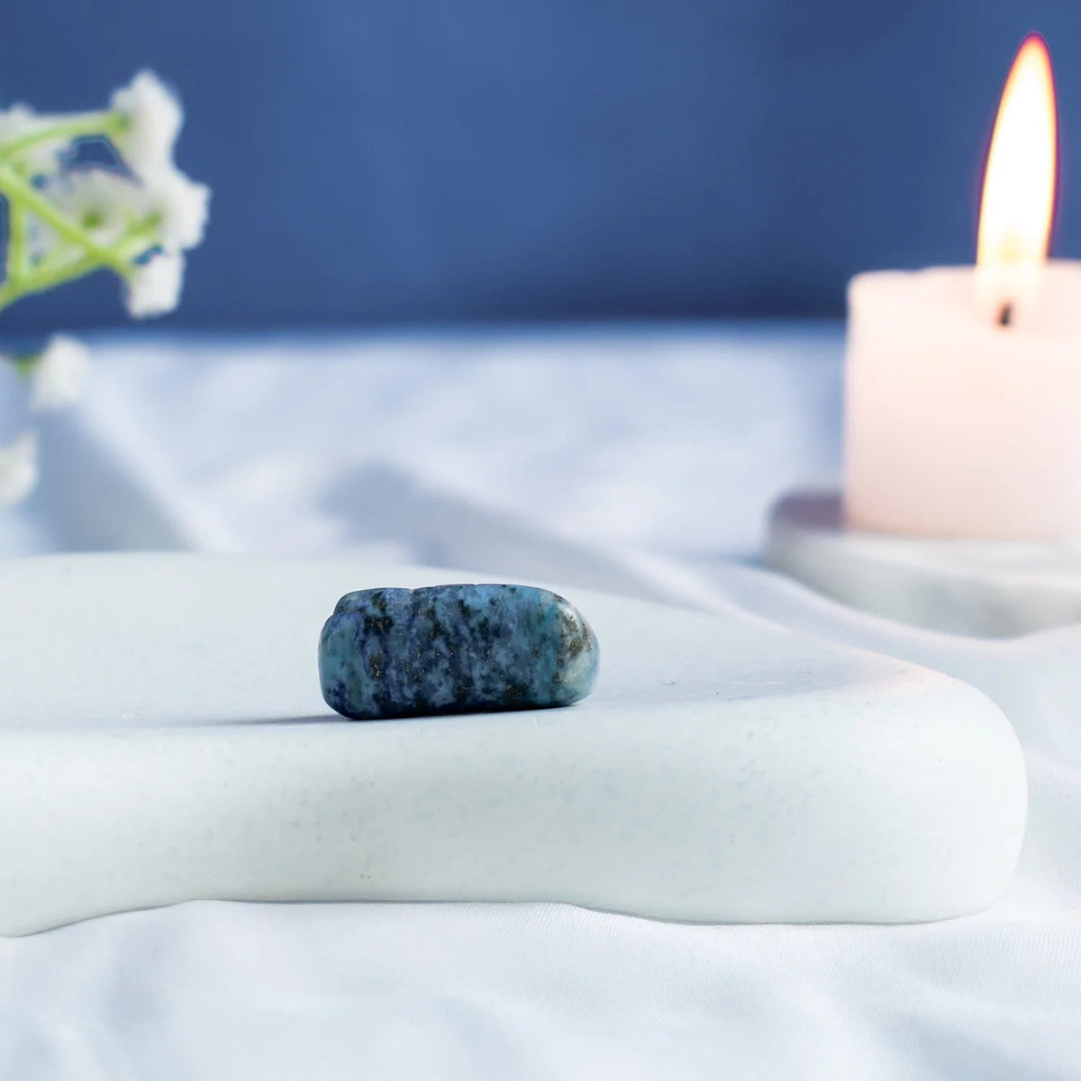 VASTUKOSHA tumbled Lapis Lazuli stone with deep blue colour and pyrite specks on a white surface with a lit candle.