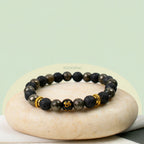 VASTUKOSHA Wealth OM bracelet featuring raw pyrite, black lava beads, and a central gold Om symbol on a stone base.