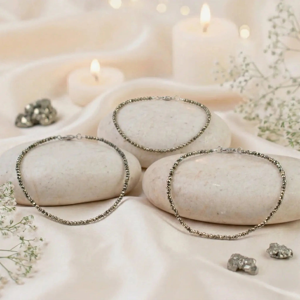 VASTUKOSHA raw pyrite anklets displayed on smooth white stones with soft candlelight and delicate baby's breath flowers.