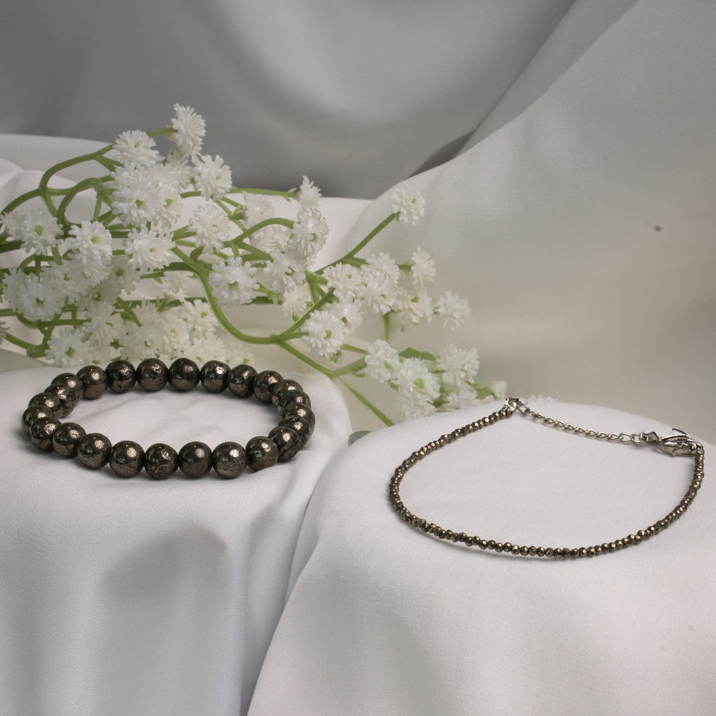 VASTUKOSHA raw pyrite bracelet and delicate pyrite anklet set displayed on white fabric with white flowers.