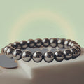 VASTUKOSHA Rinn Mukti bracelet made of polished silver hematite beads resting on a white stone plate with gold coins.