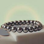 VASTUKOSHA Rinn Mukti bracelet made of polished silver hematite beads resting on a white stone plate with gold coins.